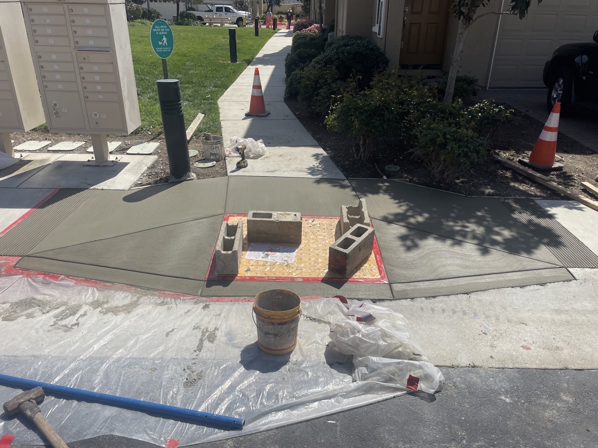 Fresh concrete at one walkway transition with covered center pad, cones and retained landscape visible